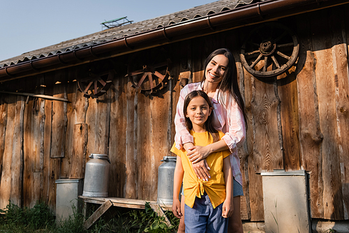 happy woman hugging daughter and smiling at camera near wooden barn