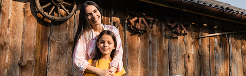 cheerful woman embracing daughter near wooden barn on farm, banner