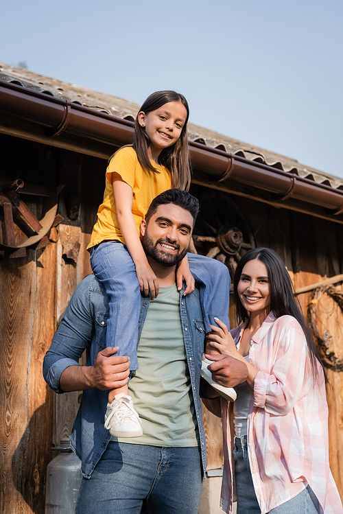 bearded man piggybacking daughter and looking at camera near happy wife on farm