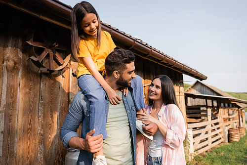 happy man piggybacking daughter near wife and wooden barn on farm