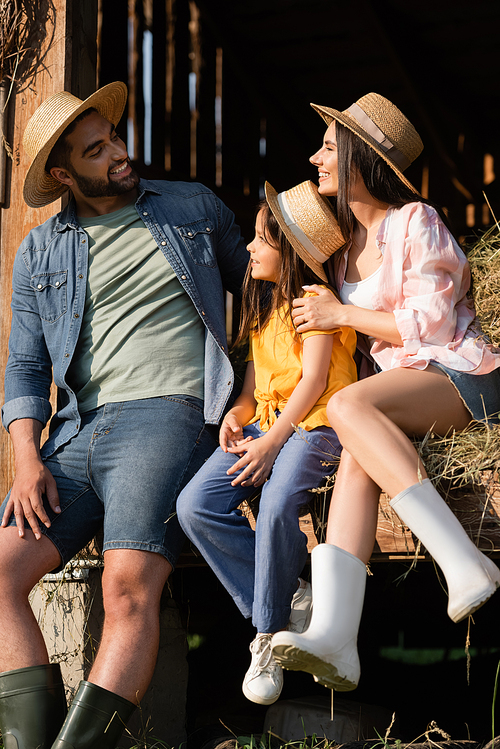 cheerful couple in straw hats looking at each other near daughter on hay in barn