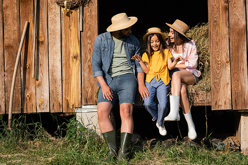 happy child in straw hat talking near parents on hay in wooden barn