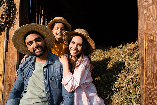 happy farm family in straw hats looking at camera near barn with hay