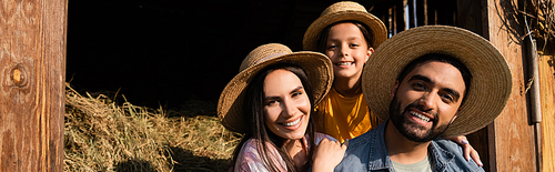 joyful farmers with daughter in straw hat smiling at camera near hay on farm, banner