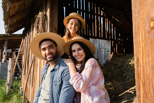 smiling village family in straw hats looking at camera near wooden barn on farm