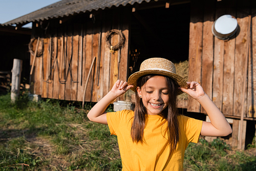 smiling child with closed eyes touching straw hat near wooden barn on blurred background