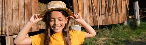 cheerful girl with closed eyes adjusting straw hat on farm in village, banner