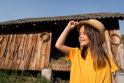happy girl in straw hat looking away near wooden barn on village farm