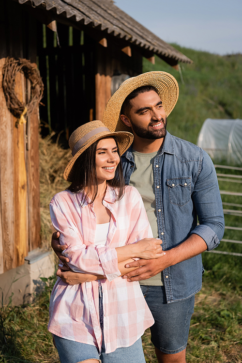 smiling couple in straw hats looking away on farm in countryside