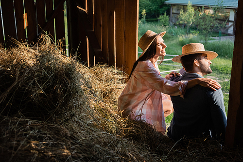 happy woman in straw hat hugging shoulders of husband while sitting on haystack in barn