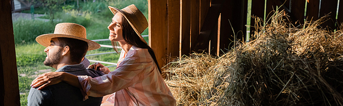 smiling woman in straw hat hugging husband near haystack in barn, banner