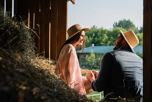 happy farmers in straw hats holding hands and looking at each other in barn near hey