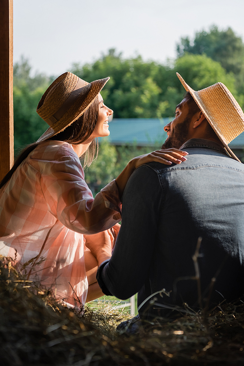 joyful woman in straw hat touching shoulder of husband near blurred hey in barn