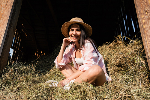 pleased woman in straw hat sitting on haystack with crossed legs and looking at camera