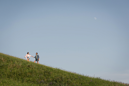 view from afar on couple holding hands and walking on green hill under blue sky