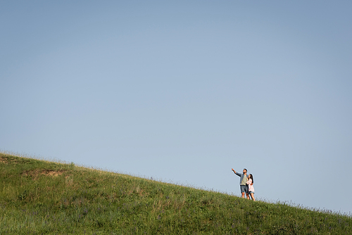 view from afar on man embracing woman and pointing with finger in green meadow