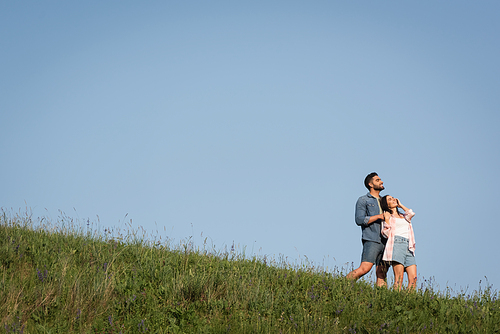 happy couple in denim shorts standing on green hill under blue sky and looking away