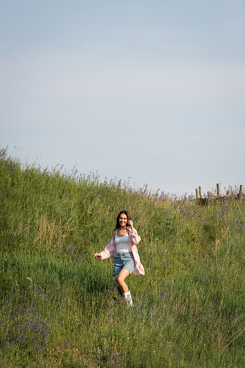 cheerful brunette woman looking at camera in green meadow with wildflowers