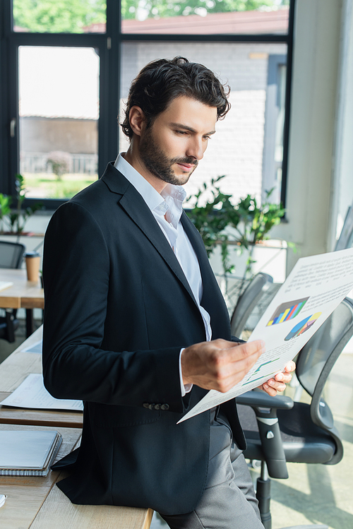 bearded businessman in black blazer looking at charts while standing in office