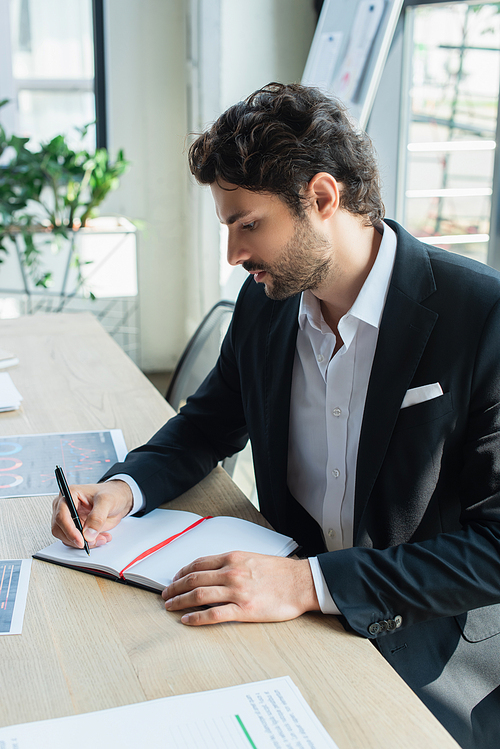 stylish businessman writing in empty notebook near papers on office desk