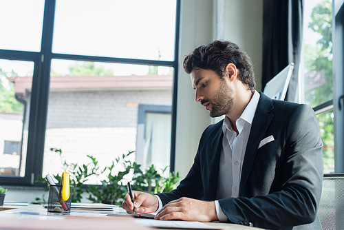 stylish businessman in black blazer writing at workplace in office