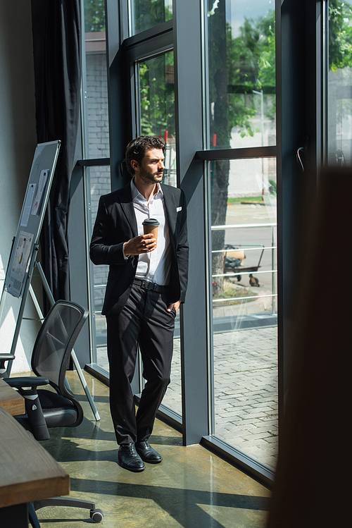 full length view of businessman in elegant suit standing with coffee to go and hand in pocket near office windows