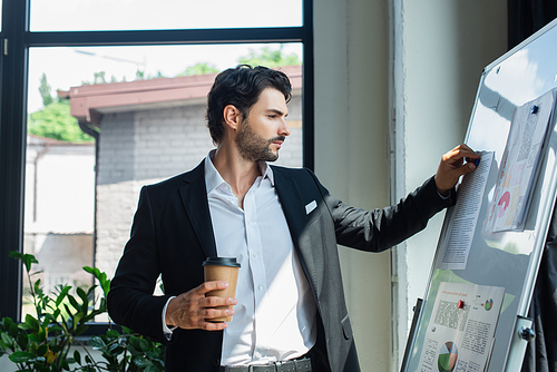 businessman in black blazer holding coffee to go near flip chart with infographics in office