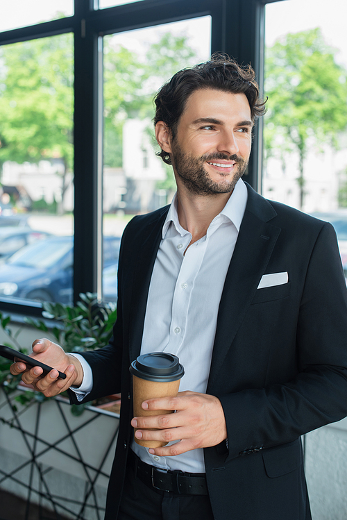 smiling businessman in black blazer holding smartphone and takeaway drink while looking away in office