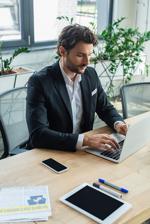 businessman in stylish formal wear typing on laptop near devices on desk