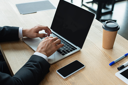 partial view of businessman typing on laptop with blank screen at workplace in office