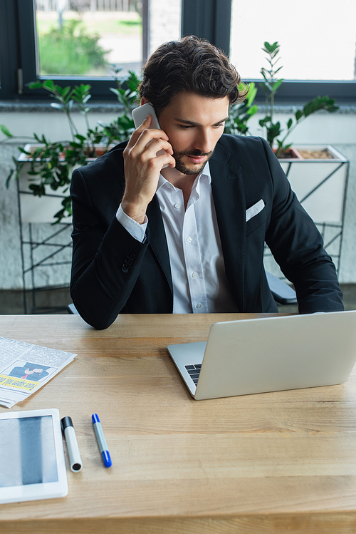 businessman in black blazer talking on smartphone near laptop at workplace