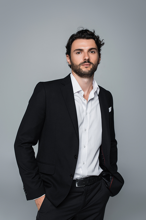 stylish man in black blazer and white shirt standing with hands in pockets isolated on grey