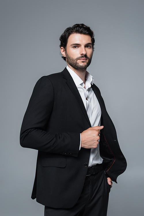 brunette man in black blazer and white shirt holding hand in pocket isolated on grey