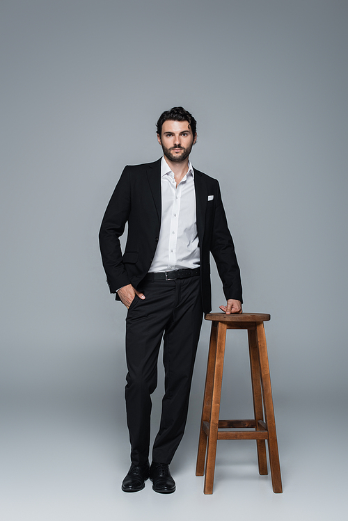 full length view of man in black suit standing with hand in pocket near high stool on grey