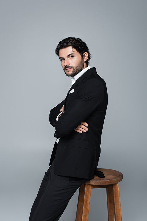 stylish man in black suit standing near high stool with crossed arms and looking at camera isolated on grey