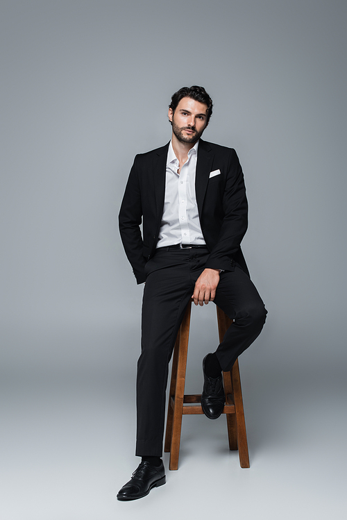 full length view of man in black suit sitting on high stool and looking at camera on grey