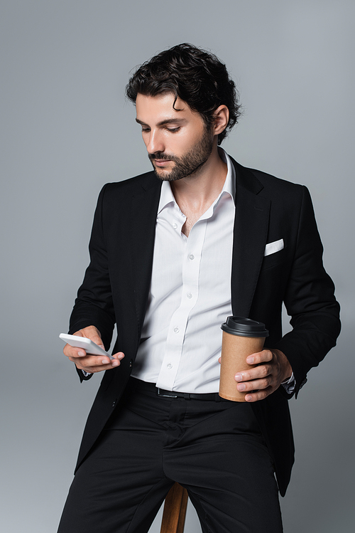 stylish brunette man with coffee to go using mobile phone while sitting isolated on grey