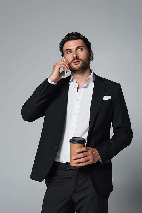 brunette man in black blazer holding coffee to go and talking on smartphone isolated on grey