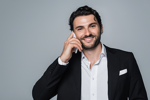 cheerful man in black blazer and white shirt talking on smartphone and looking at camera isolated on grey