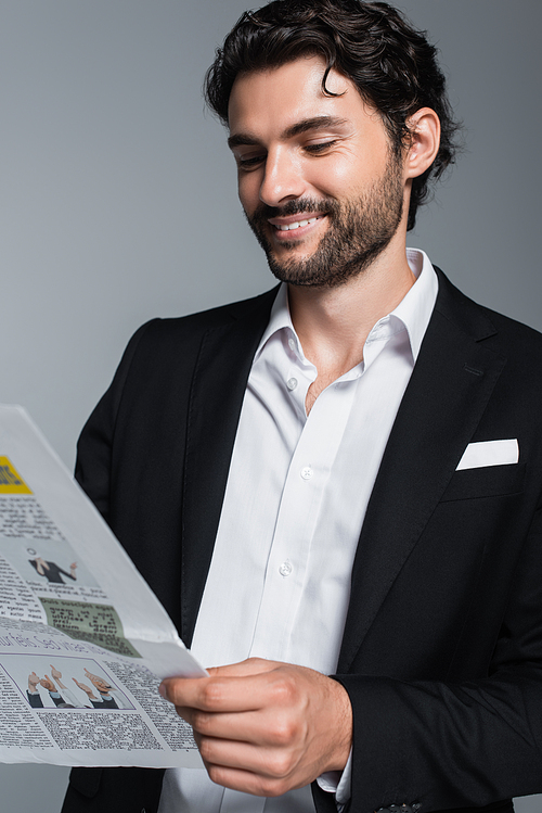 happy brunette businessman in black blazer reading newspaper isolated on grey