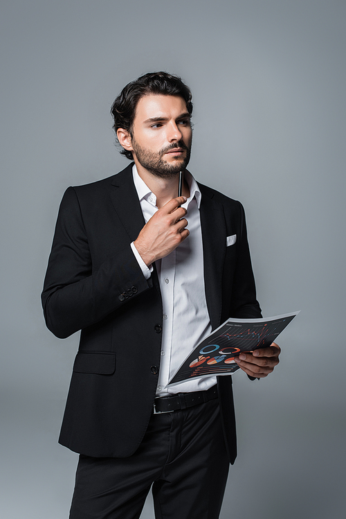 thoughtful businessman in black suit holding infographics and pen while looking away isolated on grey