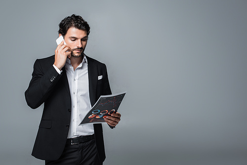 businessman in formal wear looking at charts and talking on mobile phone isolated on grey