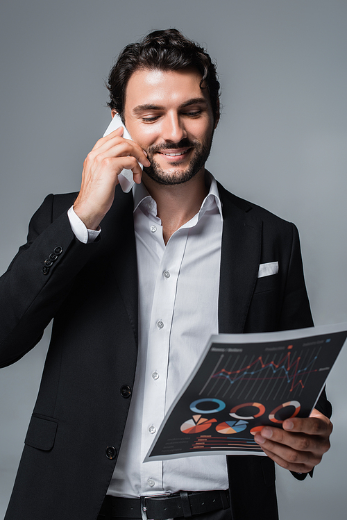 smiling businessman looking at infographics and talking on cellphone isolated on grey