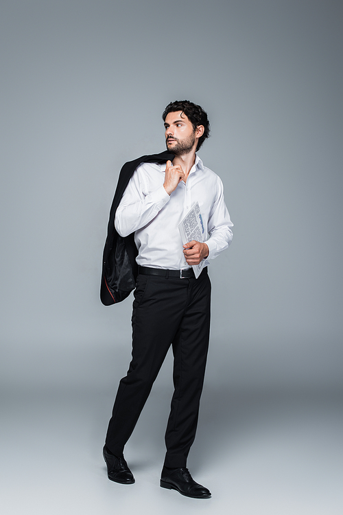 full length view of businessman holding blazer and newspaper while looking away on grey