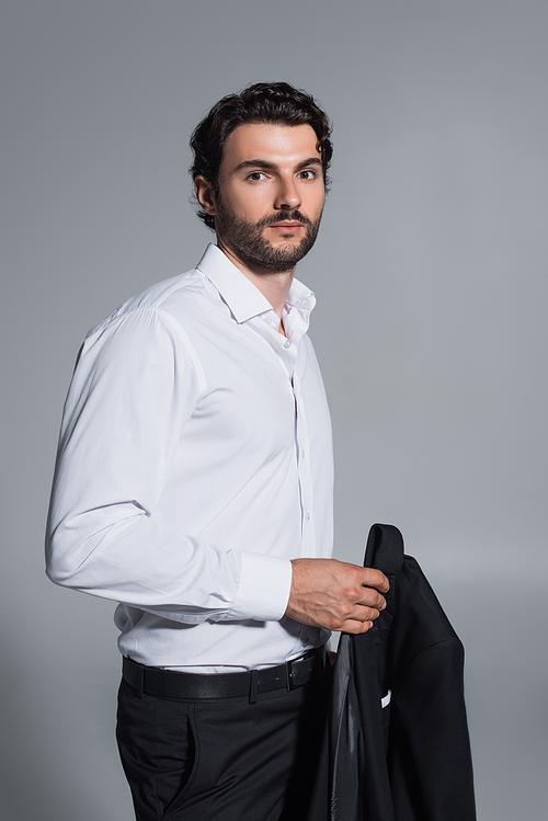 brunette man in white shirt holding black blazer while looking at camera isolated on grey