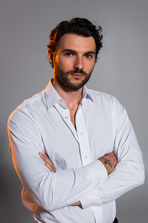 bearded man in white shirt standing with crossed arms and looking at camera on grey