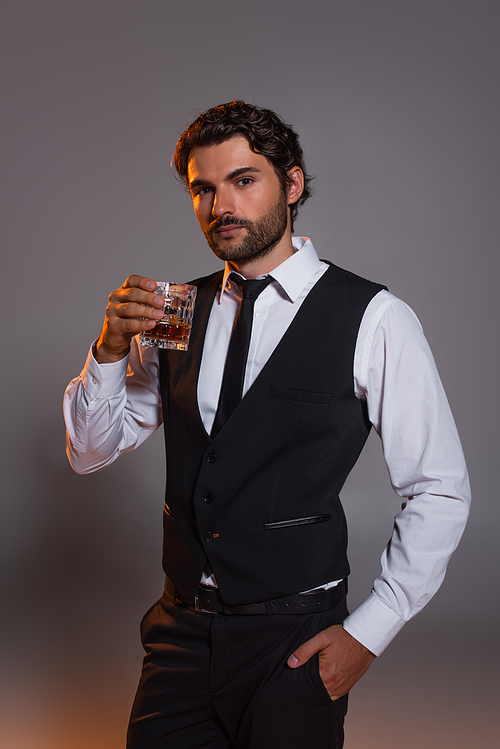 elegant brunette man with glass of whiskey standing with hand in pocket on grey background