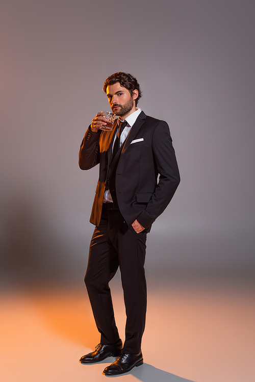 full length view of man in elegant suit holding glass of whiskey on grey background with light