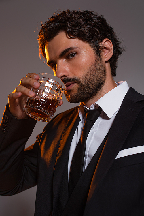 stylish man in black suit drinking whiskey while looking at camera isolated on grey