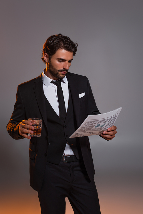 man in black elegant suit reading newspaper while holding glass of whiskey on grey background